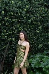 Vestido corto de lentejuelas color ciruela o morado oscuro, con escote corazón y detalle de argolla central que conecta con tirantes finos que atan al cuello. Presenta aberturas laterales (cut-outs) en la zona de la cintura y falda corta con vuelo.