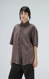 Camisa de corte oversize y mangas cortas, confeccionada en tela con efecto de teñido artesanal maltinto en tono gris. Presenta cuello clásico, cierre frontal con botones y costuras verticales decorativas en el frente.