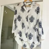 Camisa blanca de manga larga con estampado damasquinado en tono azul marino, de corte holgado y diseño asimétrico más largo en la parte trasera.