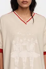 Remera oversize de color crudo con escote en V y cuello en rib rojo, mangas cortas con rib rojo y una franja roja en los hombros. Cuenta con un estampado frontal de bailarinas y detalles en relieve.