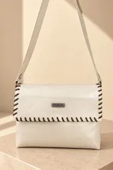 Bandolera de cuero color marfil, modelo Teka de Pecarí, con costuras cruzadas en hilo marrón oscuro en los bordes de la solapa y placa metálica frontal con la marca grabada. Incluye correa ajustable para el hombro.