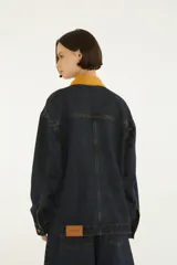 Chaqueta oversize de denim azul oscuro con cuello camisero de pana color mostaza, cierre frontal con botones metálicos y bolsillos delanteros con solapa. Cuenta con detalle de bordado en el pecho.