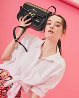 Cartera negra de cuero charolado con solapa y cierre de broche. Presenta un detalle metálico dorado en forma de freno de caballo en la parte frontal y una correa ajustable.