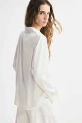 Blusa blanca de tela con brillo, cuello clásico, escote en V con botones frontales, mangas largas con puños sencillos y bajo redondeado.