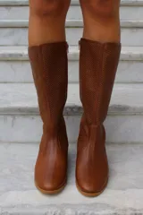 Bota de caña alta confeccionada en cuero, con diseño de textura perforada en la parte superior de la caña y puntera lisa. Cuenta con suela plana y taco bajo.