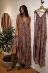 Vestido largo sin mangas, con escote en V y tirantes finos regulables con borlas en los extremos. Presenta un estampado de estilo paisley y geométrico en tonos marrones y rosados, con diseño amplio y fluido.
