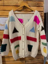 Saco de lana Merino tejido a mano con la técnica de dos agujas, de color crudo con figuras geométricas de diversos colores como rojo, azul, verde, amarillo y negro. Presenta escote en V, mangas largas y cierre frontal con botones de madera.