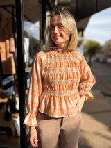 Blusa de viyela con estampado a cuadros en tonos naranja y celeste. Presenta un diseño con frunces en el cuerpo, cuello alto, mangas largas abullonadas con puños elásticos y terminación con volados.