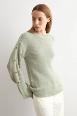 Sweater de punto color blanco natural, con cuello redondo y mangas largas adornadas con botones dorados a lo largo de las costuras exteriores.
