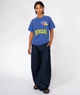 Remera de algodón gris oscuro con lavado especial, corte holgado y cuello redondo. Presenta un estampado frontal con la palabra "MICHIGAN" en amarillo neón con borde negro, y apliques decorativos en la parte superior: un corazón naranja, la palabra "NEWTONE" en arco y dos píldoras/pastillas de colores.