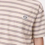 Remera de manga corta con diseño a rayas horizontales en tonos beige, marrón y gris. Presenta un bolsillo frontal en el pecho con el logo de la marca bordado y un corte holgado.