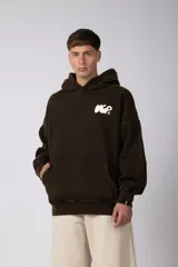 Buzo canguro oversize de algodón frisa premium con efecto lavado, color marrón oscuro. Presenta capucha, bolsillo frontal tipo canguro, puños y ruedo acanalados, y logo estampado en el pecho.
