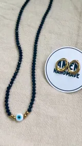 Conjunto de joyería que incluye un choker de cristal checo negro con dije de ojo turco central y detalles dorados, acompañado de aros abridores de acero quirúrgico con diseño orgánico.