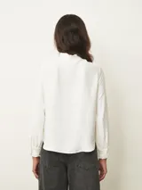 Camisa de manga larga en color blanco, con cuello mao y detalle de volados sutiles en el borde. Presenta cierre frontal oculto con botones y puños con volados.