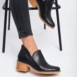 Botín de cuero vacuno color negro, con diseño de corte lateral, cierre metálico en el talón y taco foliado de madera en tono natural.