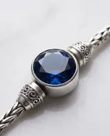 Pulsera de plata con diseño trenzado y una piedra circonia redonda facetada en color azul zafiro en el centro.
