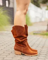 Bota estilo texana de caña alta, confeccionada en cuero gamuzado color marrón óxido. Presenta un diseño de corte en V en la parte superior de la caña, costuras decorativas y un taco bajo de madera.