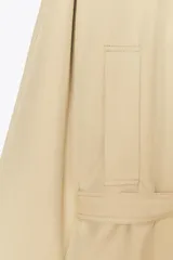 Trench color beige con cuello de solapa subido y manga larga. Tiene bolsillos laterales, forro interior y cinturón de lazo en el mismo tejido. El cuello se cierra con un botón.