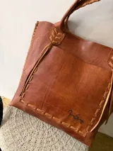 Cartera tipo shopper de cuero genuino marrón, con terminaciones artesanales trenzadas en las asas y los laterales, bolsillo frontal externo y asas largas para llevar al hombro.