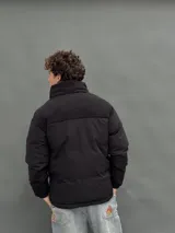 Campera puffer negra con diseño de paneles, cierre frontal completo y cuello alto. Presenta una textura diferenciada en la parte superior y bolsillos laterales.