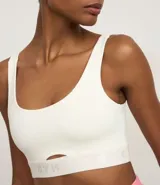 Top deportivo color blanco hueso, de corte ajustado, con breteles anchos y banda elástica inferior con inscripción. Presenta un pequeño recorte circular en el centro del busto.