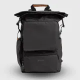 Mochila urbana de diseño funcional en color mostaza, confeccionada en material repelente al agua. Cuenta con cierre superior enrollable con hebillas ajustables, un compartimento frontal grande con cierre, un bolsillo inferior adicional con el logo de la marca, compartimentos internos organizadores, funda acolchada para laptop y bolsillos laterales para botellas.