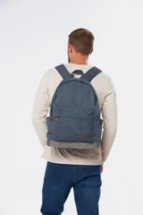 Mochila de lona tipo canvas en color azul con base en tono grisáceo y detalles en cuero sintético marrón. Cuenta con un compartimento principal amplio, un bolsillo frontal con cierre, bolsillos laterales abiertos y tirantes ajustables.