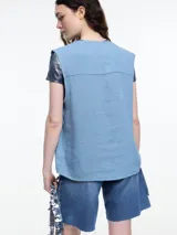 Chaleco sin mangas color coral, con escote en V, cierre frontal con botones de madera y bolsillos de parche. Incluye tres pins decorativos: un corazón plateado, un círculo plateado y un cuadrado rojo. Confeccionado en tejido liviano de lino y viscosa.