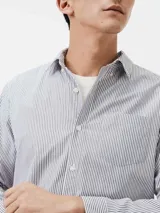 Camisa de manga larga con cierre frontal de botones, cuello clásico y bolsillo en el pecho. Presenta un diseño de rayas verticales finas en tonos azul y blanco.