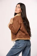 Campera tipo gamulán de gamuza color tostado con interior y solapas de corderito sintético color crema. Presenta cierre frontal con botones, bolsillos cuadrados delanteros y detalles de costuras visibles.