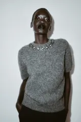 Jersey de punto de manga corta en color gris jaspeado, con cuello redondo decorado con abalorios joya y cierre de botón en la espalda.