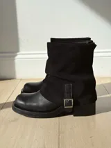 Botas de caña media combinadas en cuero liso y gamuza negra, con diseño de solapa superior y detalle de tira con hebilla metálica lateral.