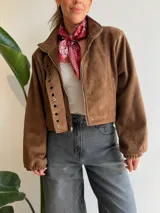 Chaqueta corta de efecto gamuza con cierre frontal y botones. Presenta mangas tipo globo con puños elásticos y detalle de aros metálicos decorativos en la solapa. Cuenta con ajuste lateral en la cintura y forro interior.