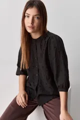 Camisa negra de textura liviana con detalle de lunares en relieve, cuello mao y cierre frontal con botones. Presenta mangas 3/4 con puños elastizados y frunces en los hombros.