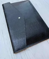 Funda para laptop de diseño minimalista, confeccionada en cuero negro con textura grabada tipo reptil y cierre mediante botón metálico a presión.