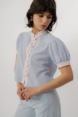 Blusa celeste a rayas blancas, de manga corta abullonada. Cuenta con cuello mao y tapeta de botones con detalles de estampado floreado en tono rosado, presentes también en el cuello y los puños.