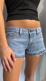 Short de jean tiro bajo en color azul claro, con diseño de cinco bolsillos y costuras reforzadas en color rojo. Presenta un ruedo deshilachado con un detalle de costura horizontal que le otorga un aspecto desgastado.