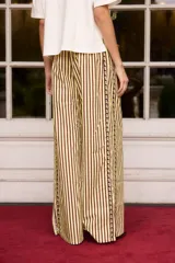 Pantalón de corte recto y tiro alto, confeccionado en tela de algodón con diseño de rayas verticales en tonos oscuros. Presenta una banda lateral decorativa con pequeños detalles bordados en color amarillo.