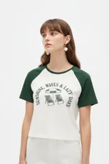 Remera de manga corta estilo raglán, de color blanco con mangas y cuello redondo en verde oscuro. Presenta un estampado frontal con texto y un diseño gráfico de reposeras y sombrilla de playa.