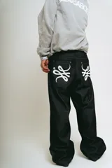 Pantalón de jean negro, corte baggy, confeccionado en denim crudo. Presenta un estampado serigrafiado en blanco con un diseño estilizado en ambos bolsillos traseros y un parche con logo en la cintura.