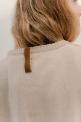 Buzo de lambswool color beige, con escote redondo y detalle en V. Presenta cierres metálicos en los puños que revelan un interior en contraste de color marrón y ribete rojo. Tiene tajos laterales para mayor comodidad y un fit amplio.