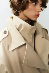 Trench corto oversize color beige con cuello subido de efecto doble, cierre cruzado con botones oscuros y bolsillos de vivo delanteros.
