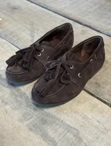 Mocasín náutico de gamuza color marrón chocolate, con cordones decorativos, detalle de flecos y borlas en el empeine, y ojales metálicos laterales.