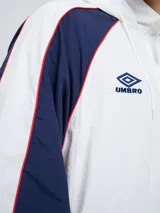 Campera deportiva Umbro del Club Nacional de Football, modelo Venti, para hombre. Diseño en color blanco con mangas azules y detalles de líneas rojas. Cuenta con capucha con cordón ajustable, cierre frontal y bolsillos. Presenta el logo de Umbro y el escudo del club bordados en el pecho.