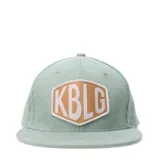 Gorro tipo cap de pana color verde agua, con parche frontal hexagonal en color beige y letras blancas con la inscripción KBLG.
