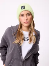 Gorro tipo beanie de punto texturizado en color verde claro, con parche rectangular negro con la inscripción 'EAT THE WAVE' en la parte frontal.