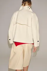 Trench corto color marfil de algodón, con cuello camisero, solapa cortaventos, mangas largas amplias con puños abotonados y cierre cruzado con un botón visible.