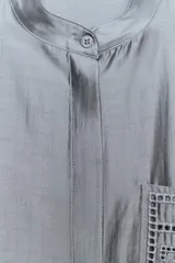 Camisa de cuello mao y manga larga con puños anchos, de color gris satinado. Cuenta con un bolsillo de plastrón en el pecho con detalles bordados a tono, al igual que los puños, y cierre frontal con botones ocultos bajo solapa.