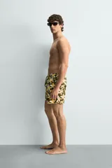 Bañador de hombre con estampado floral en tonos amarillo y blanco sobre fondo verde oliva. Presenta cintura elástica ajustable con cordón, bolsillos frontales y forro interior.