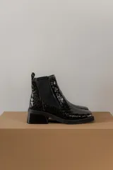 Botas tipo chelsea de cuero color marrón oscuro con acabado brillante. Presentan punta cuadrada con detalle de puntera metálica, elásticos laterales y taco cuadrado bajo.
